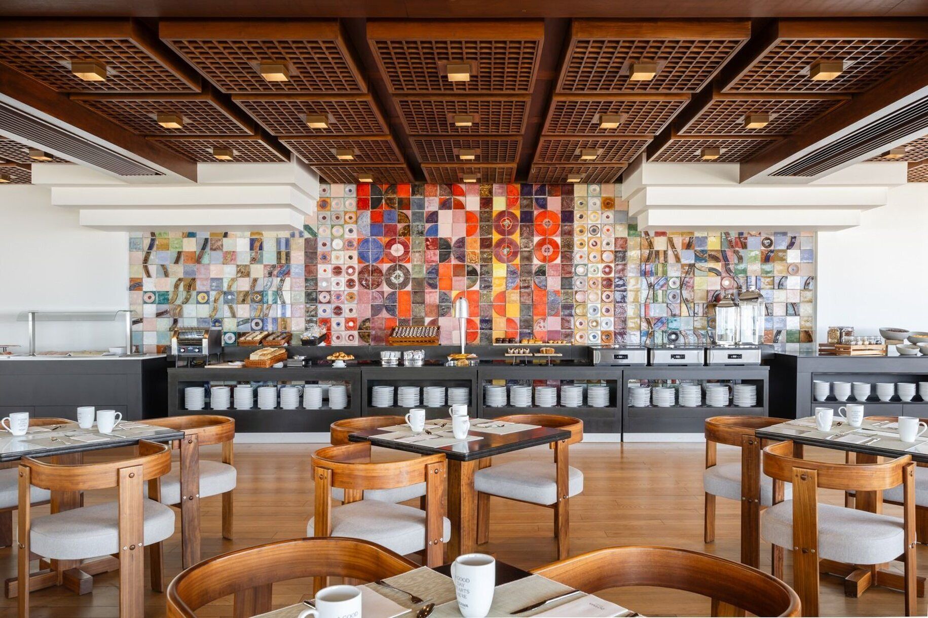 Un comedor moderno con mesas y sillas de madera, un mostrador de buffet con diferentes alimentos y una colorida instalación artística en la pared.