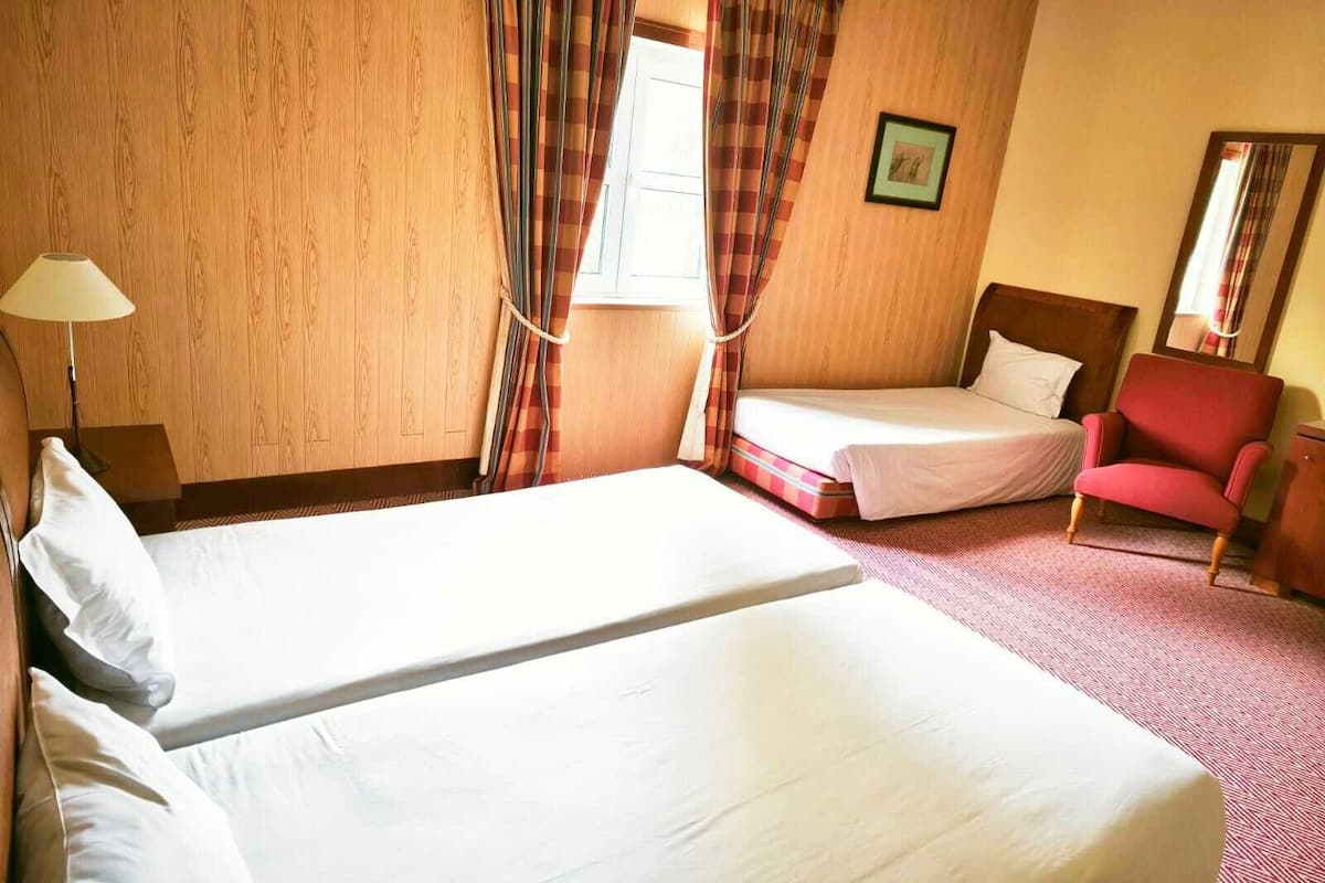 A hotel room with two beds, a red armchair, a bedside table, a lamp, and a window with curtains.