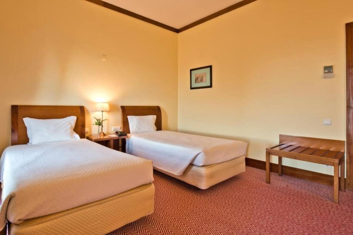 A hotel room with two beds, a bench, a nightstand with a lamp, and a framed picture on the wall.