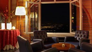 A cozy seating area with a table and chairs, a lamp, and a view of a cityscape at night. - Lam Hotel Caramulo  (Tondela)