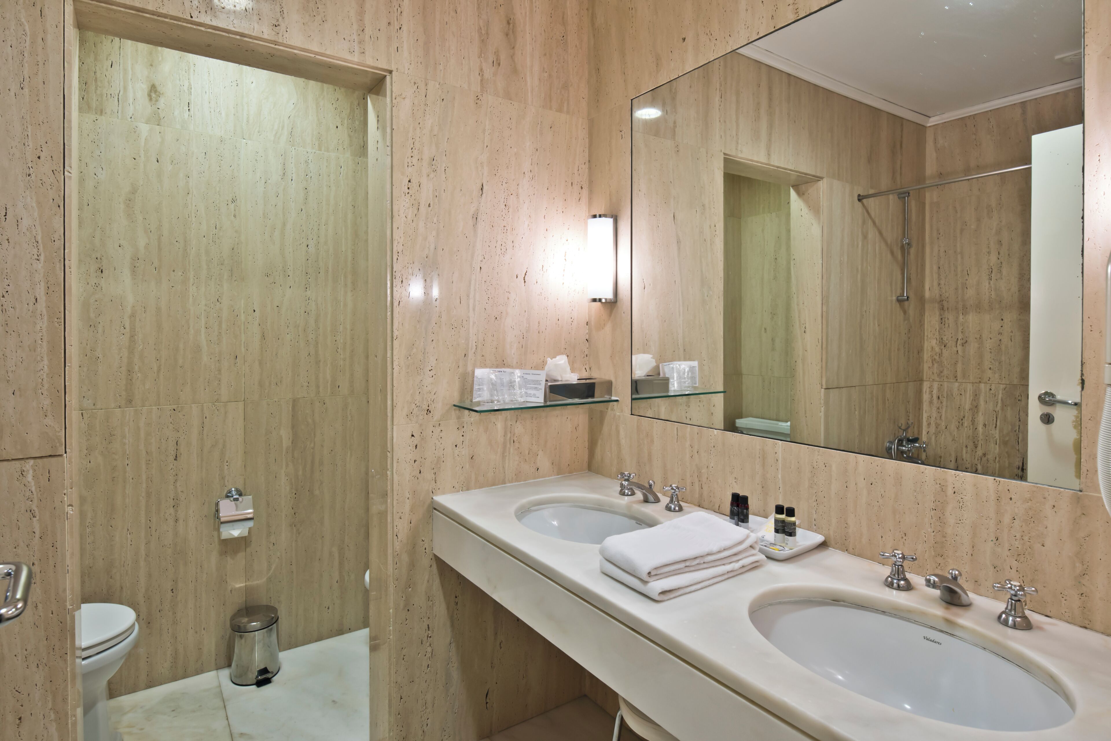 Un baño moderno con dos lavamanos, un inodoro, un estante de vidrio con artículos de aseo y un espejo grande.