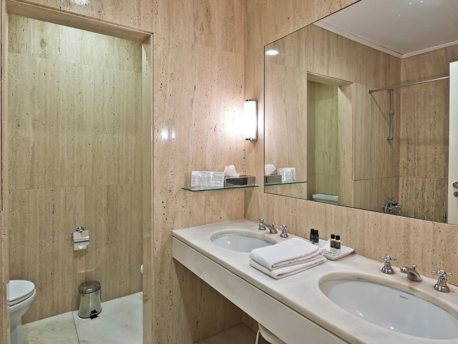 Un baño moderno con dos lavamanos, un inodoro, un estante de vidrio con artículos de aseo y un espejo grande.