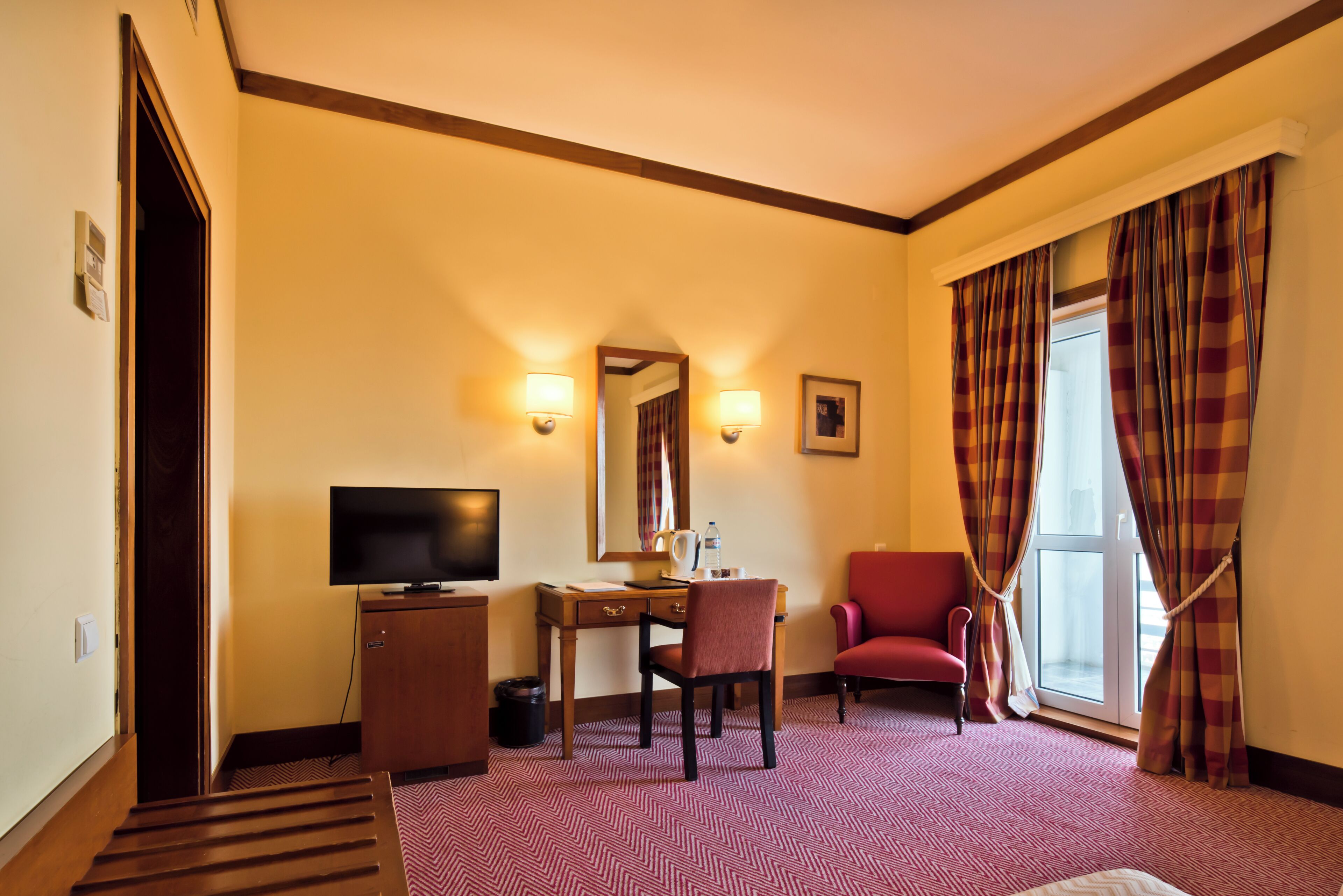 A hotel room with a desk, chair, television, and a red armchair.