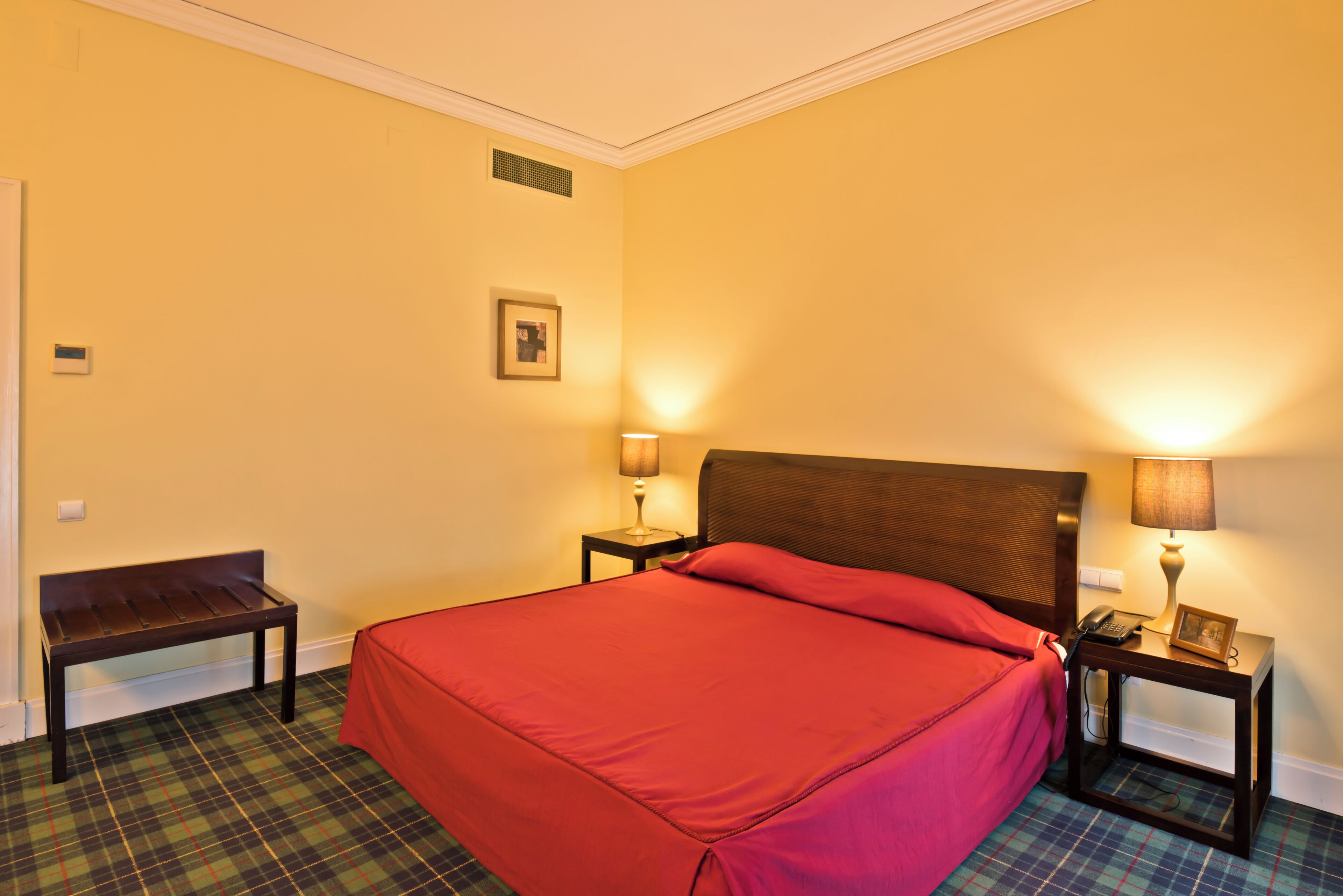 A hotel room with a bed, two bedside tables, a bench, a wall-mounted picture, and a vent.