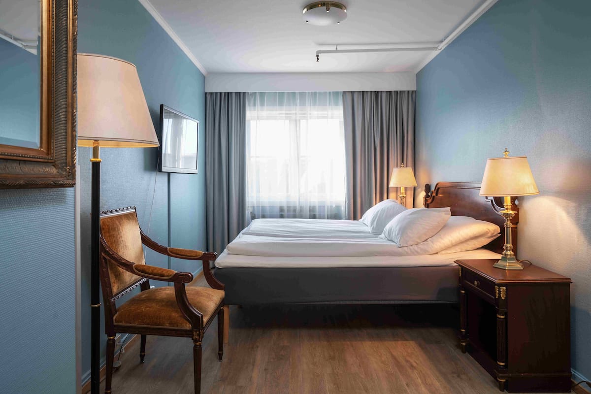 A hotel room with a bed, a chair, a nightstand with a lamp, and a window with curtains.