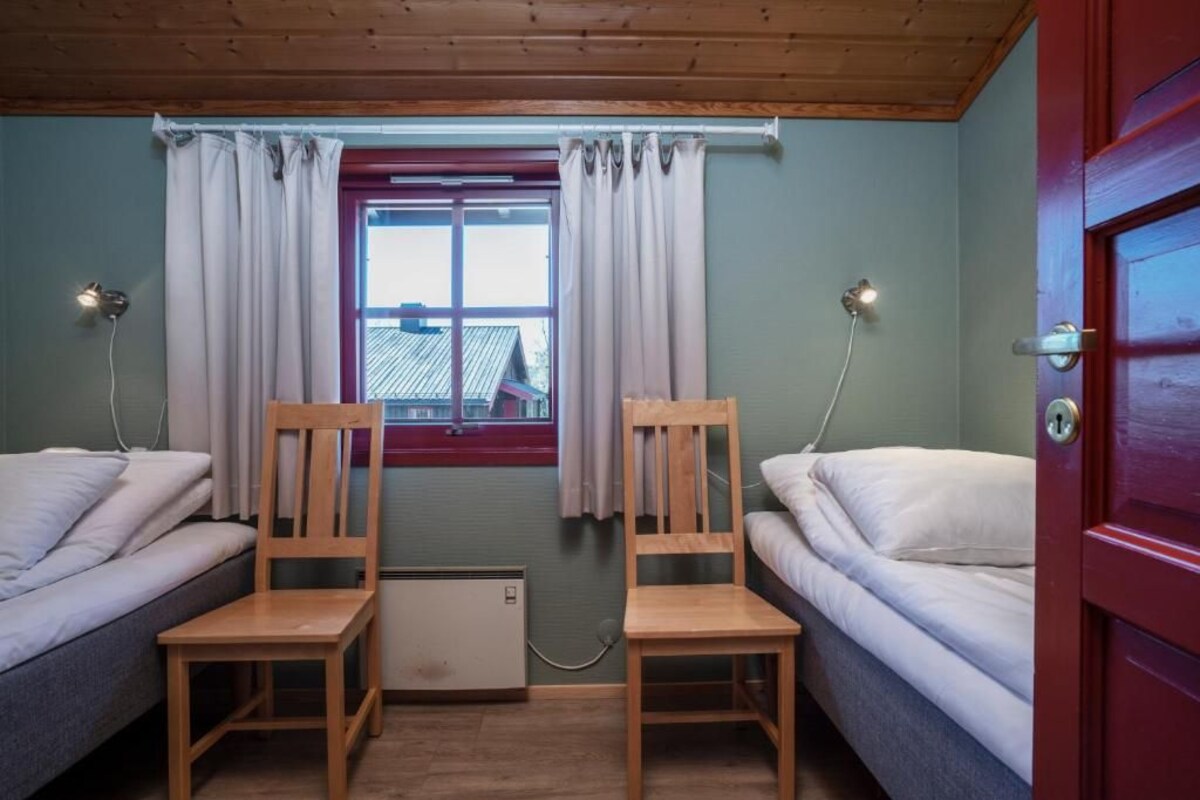 A room with two single beds, wooden chairs, a radiator, a window with curtains, and a red door.