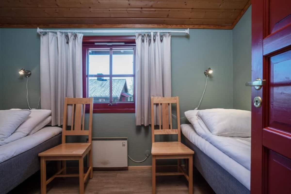 A room with two single beds, wooden chairs, a radiator, a window with curtains, and a red door.