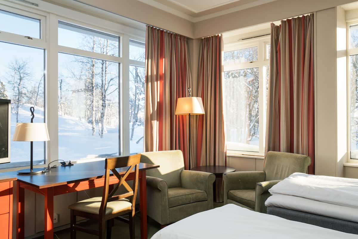 A hotel room with a desk, two chairs, a bed, and large windows with curtains.