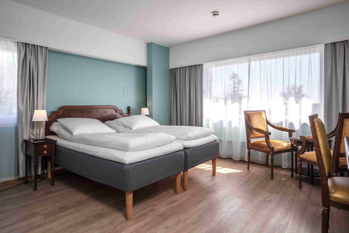 Une chambre d’hôtel avec un lit double, deux tables de chevet avec des lampes, une chaise en bois et une grande fenêtre avec des rideaux.