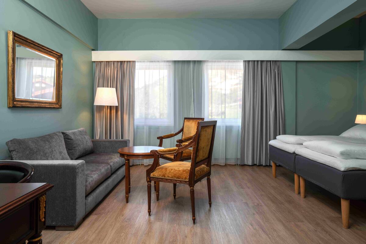 Une chambre d’hôtel avec un canapé gris, une chaise en bois, une petite table, un lit et une fenêtre avec des rideaux.