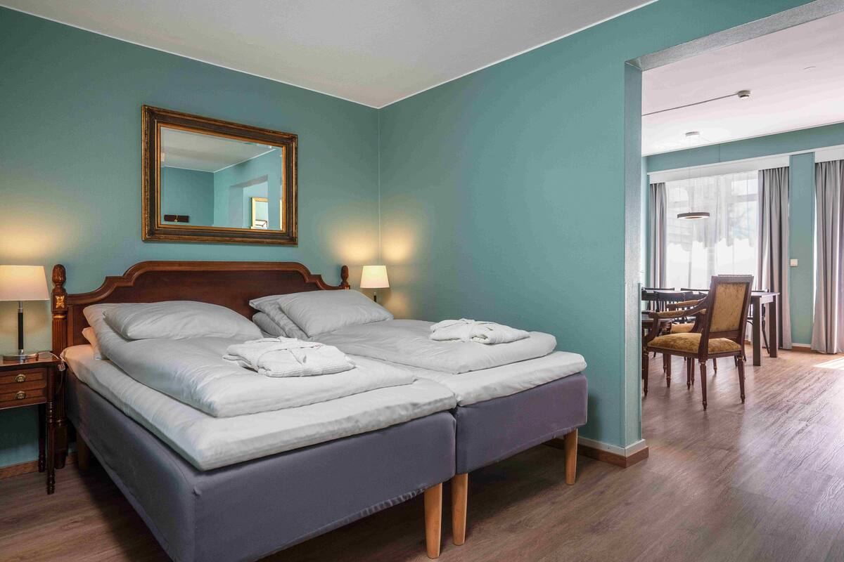 Un lit double avec une tête de lit en bois, deux tables de chevet avec des lampes, un miroir au-dessus du lit et une vue sur un coin repas avec une table et des chaises.