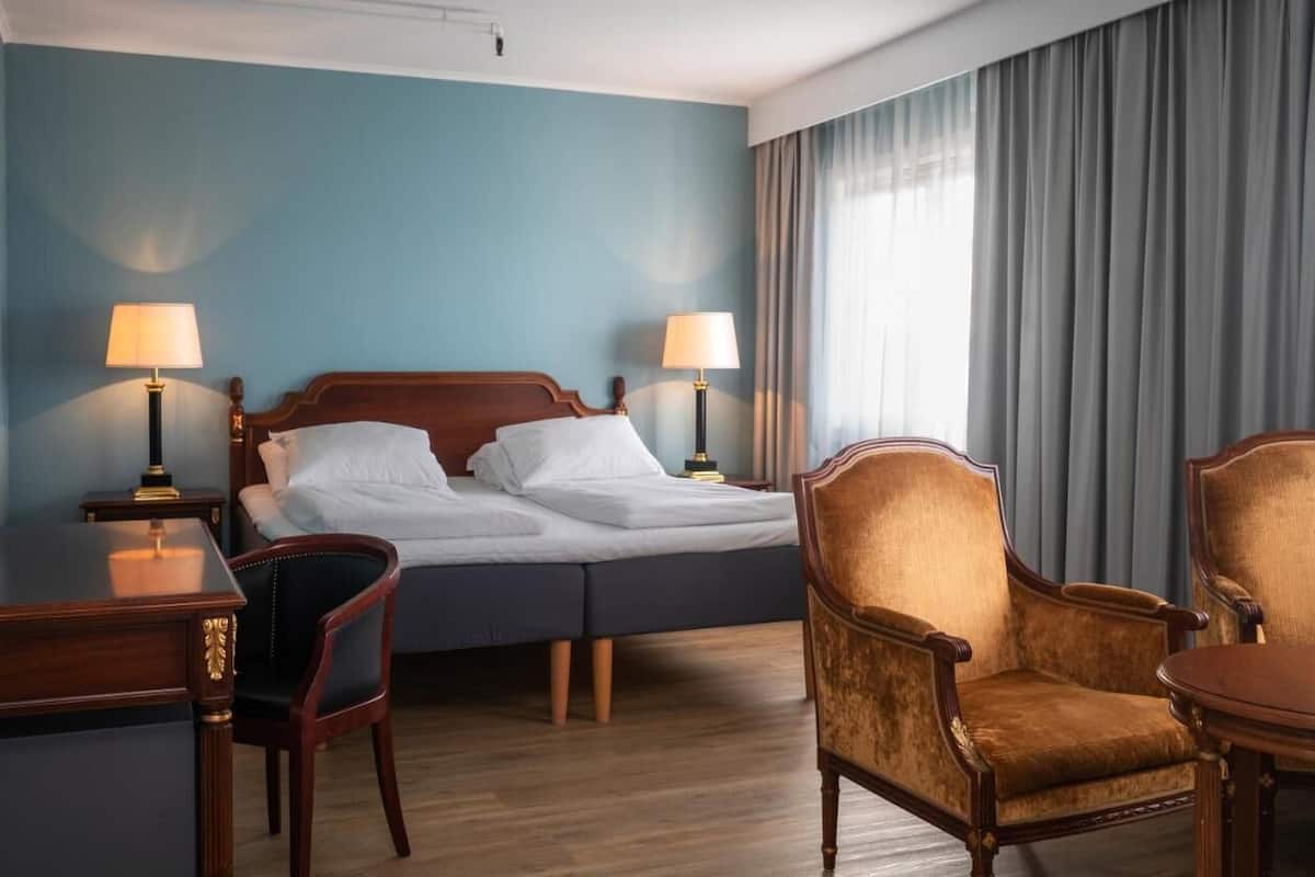 A hotel room with a double bed, two bedside lamps, a desk, a chair, and a dining table.