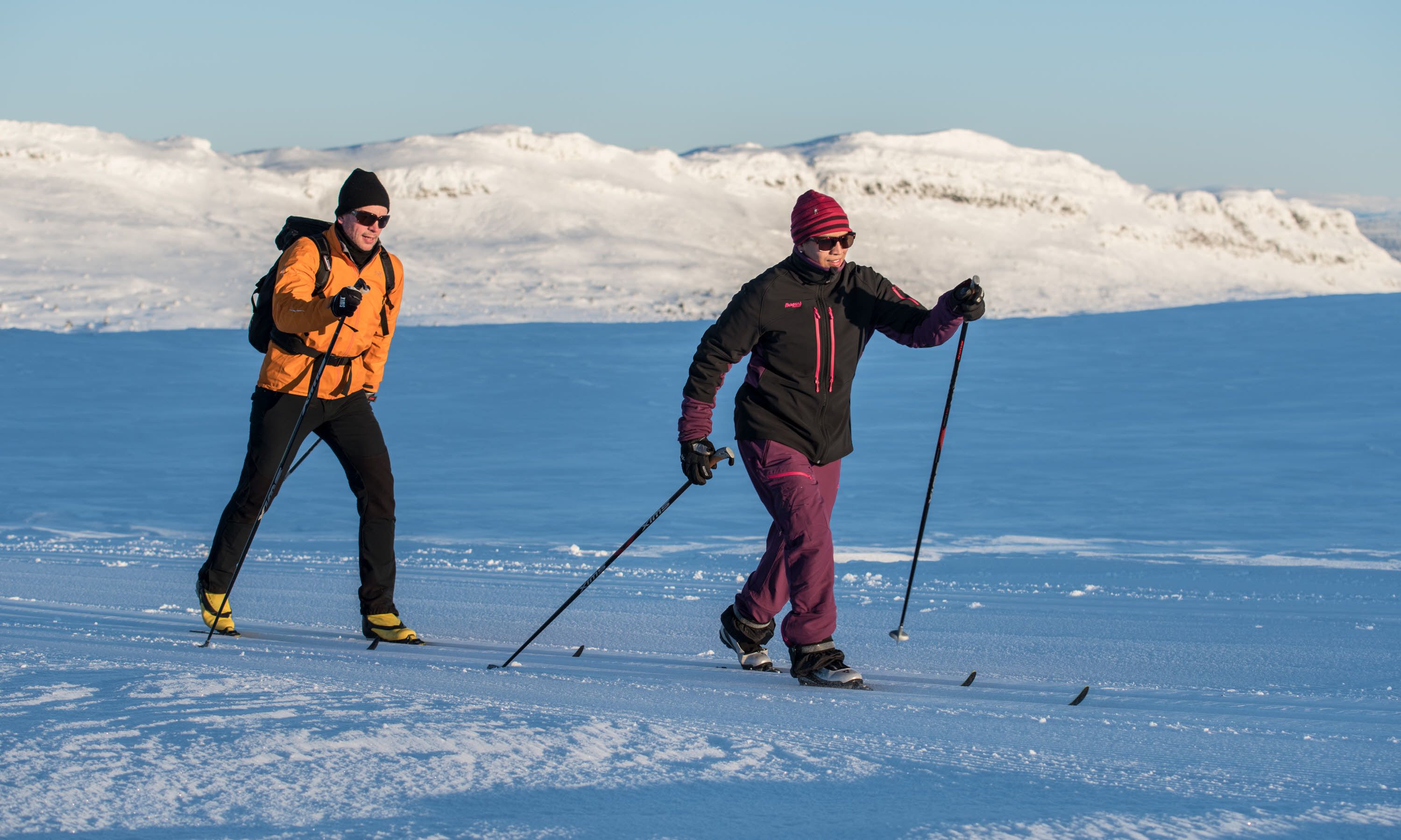 Two people cross-country skiing on a snowy landscape with mountains in the background.