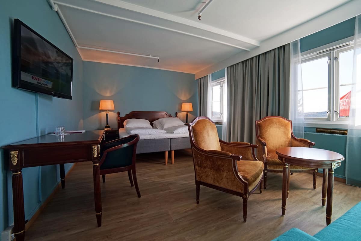 A hotel room with a bed, two armchairs, a small table, a desk, and a TV mounted on the wall.