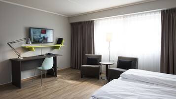 Habitación de hotel con cama, escritorio con silla, una mesita redonda con una silla, televisión y lámpara.