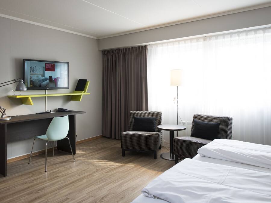 Habitación de hotel con cama, escritorio con silla, una mesita redonda con una silla, televisión y lámpara.