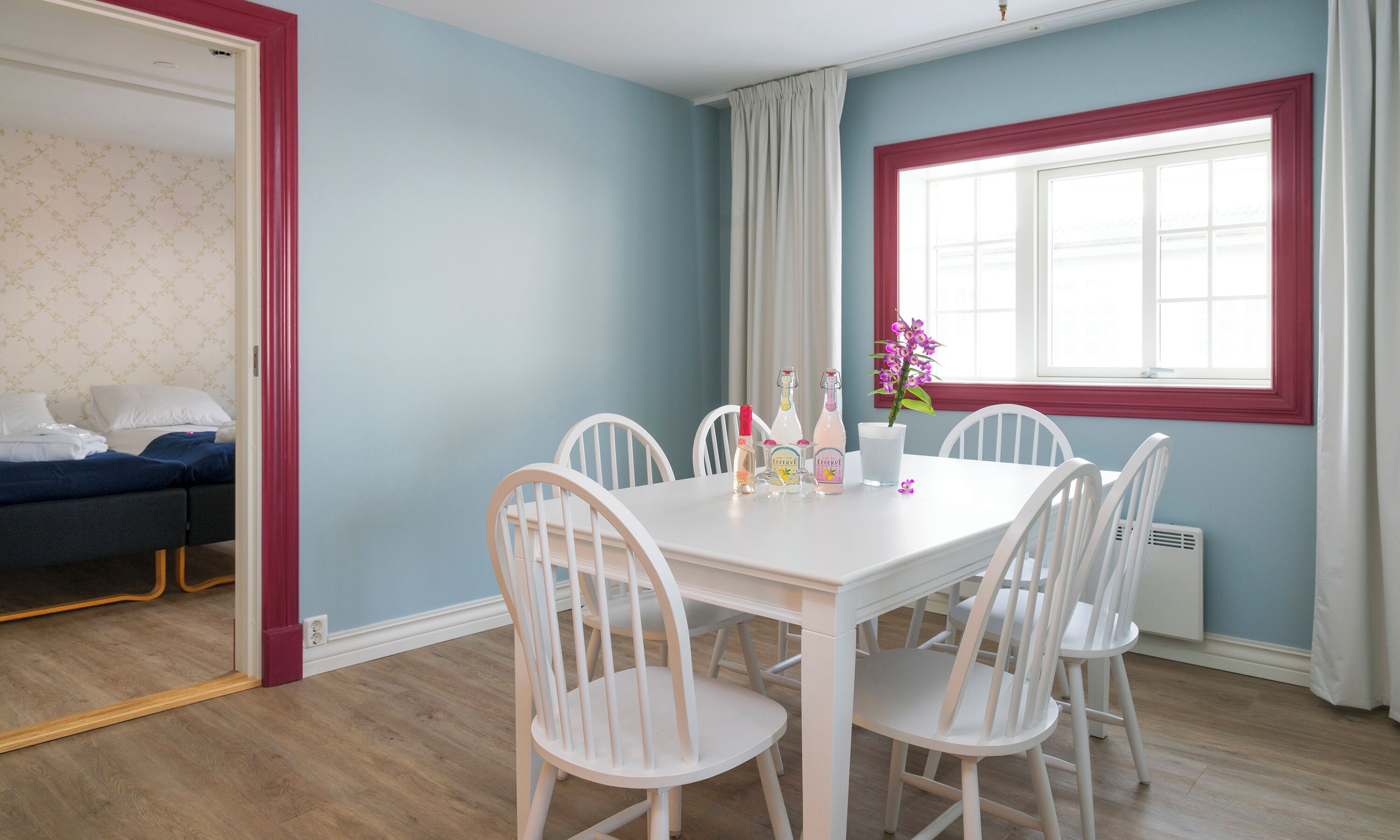Une pièce comprenant une table à manger, des chaises blanches, une fenêtre avec des rideaux et un lit avec une tête de lit bleue.