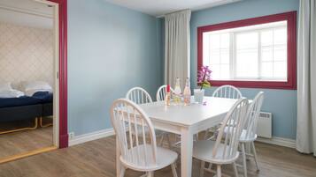 A room with a dining table, white chairs, a window with curtains, and a bed with a blue headboard.