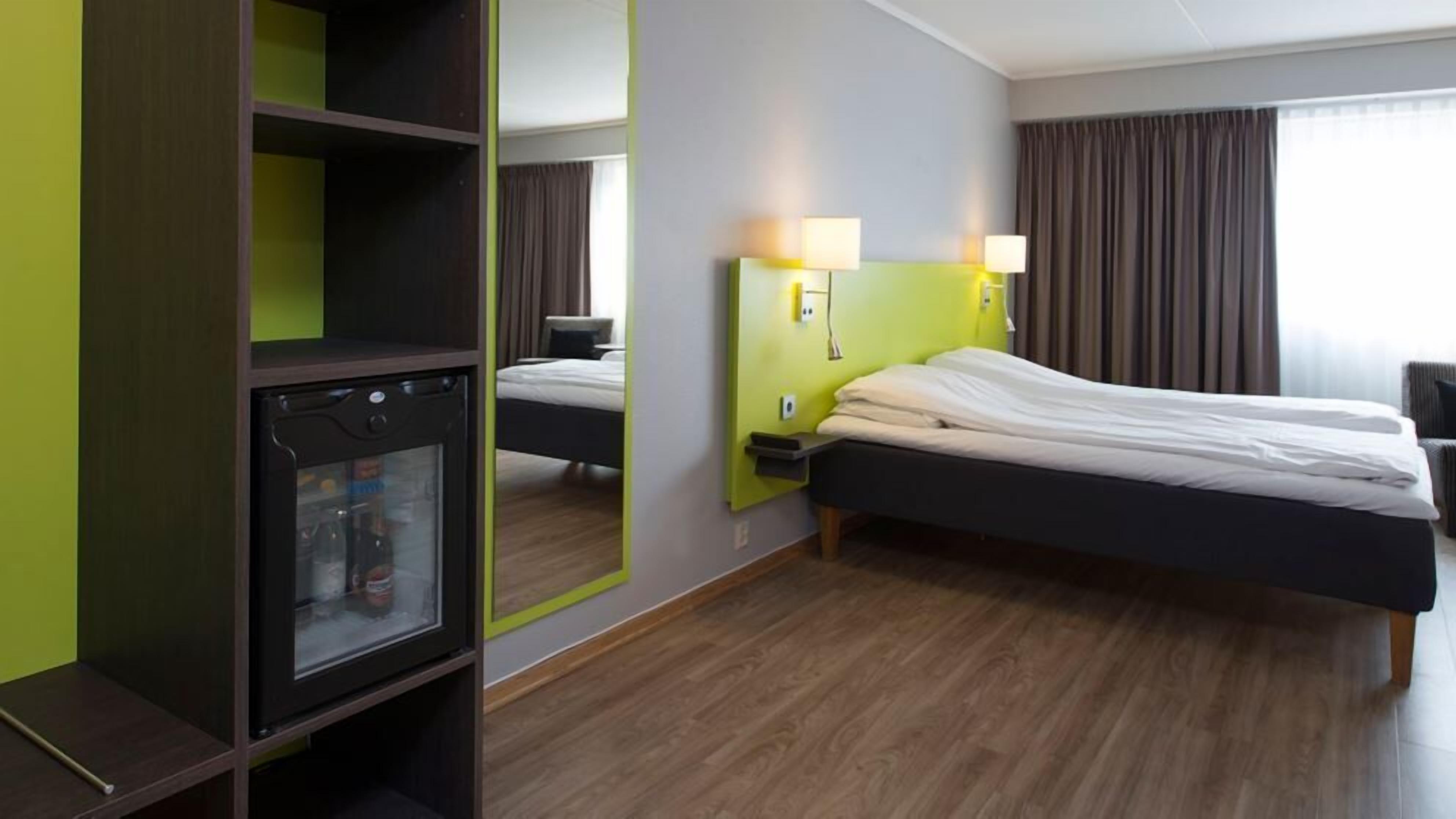 Habitación de hotel con cama, mesita de noche, lámpara, armario y minibar.