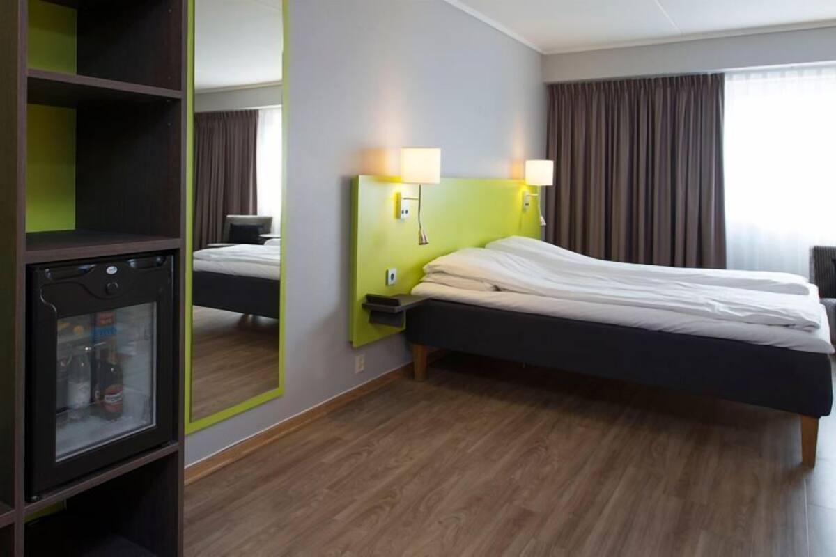 A hotel room with a bed, a bedside table, a lamp, a wardrobe, and a minibar.