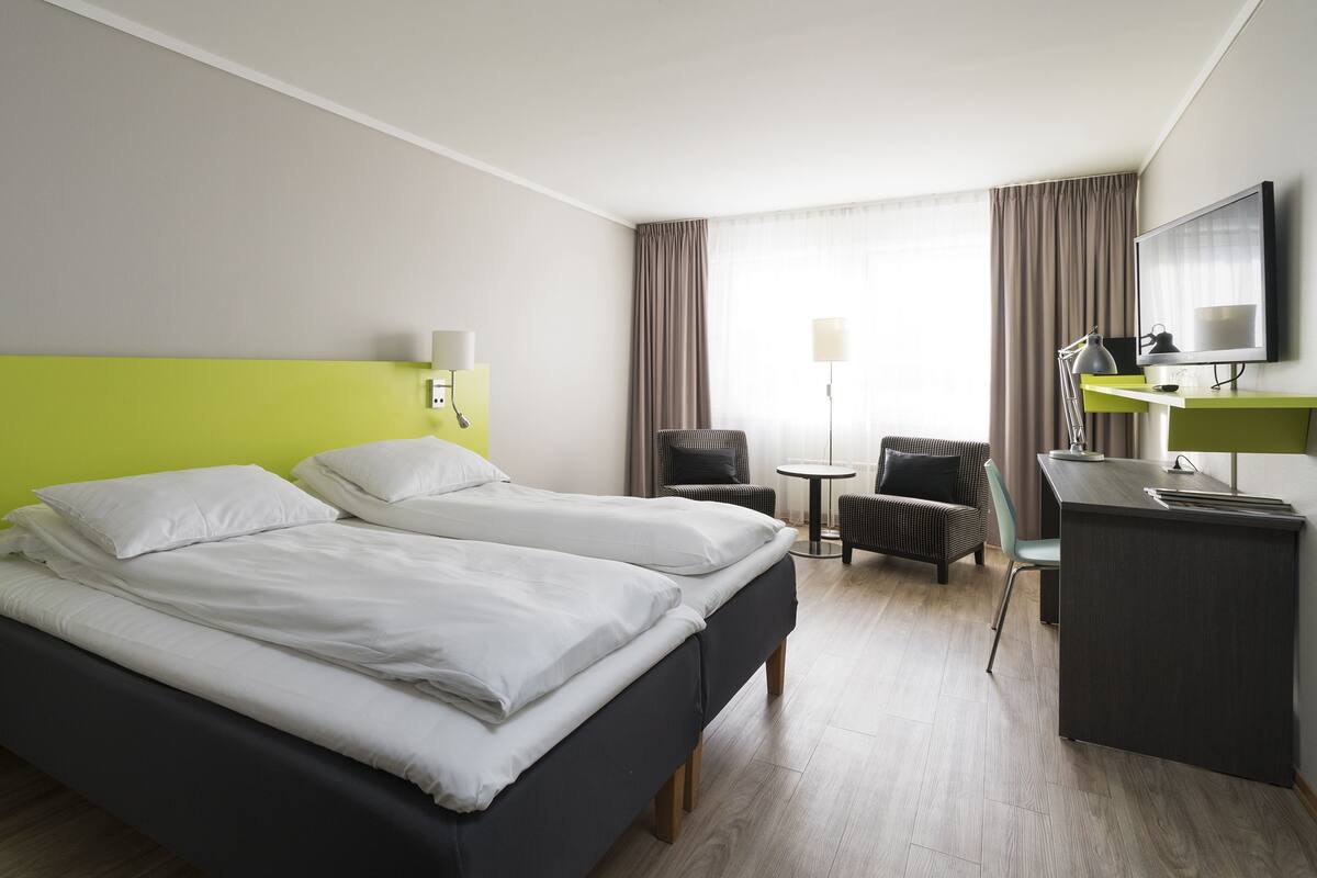 A hotel room with two beds, a desk, a chair, a TV, and a lamp.
