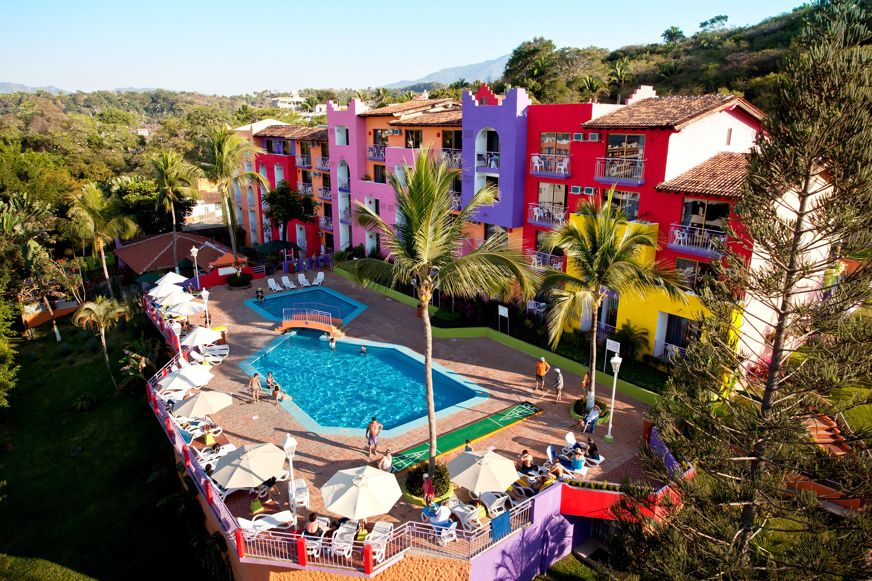A colorful hotel with a swimming pool surrounded by palm trees and lounge chairs.
