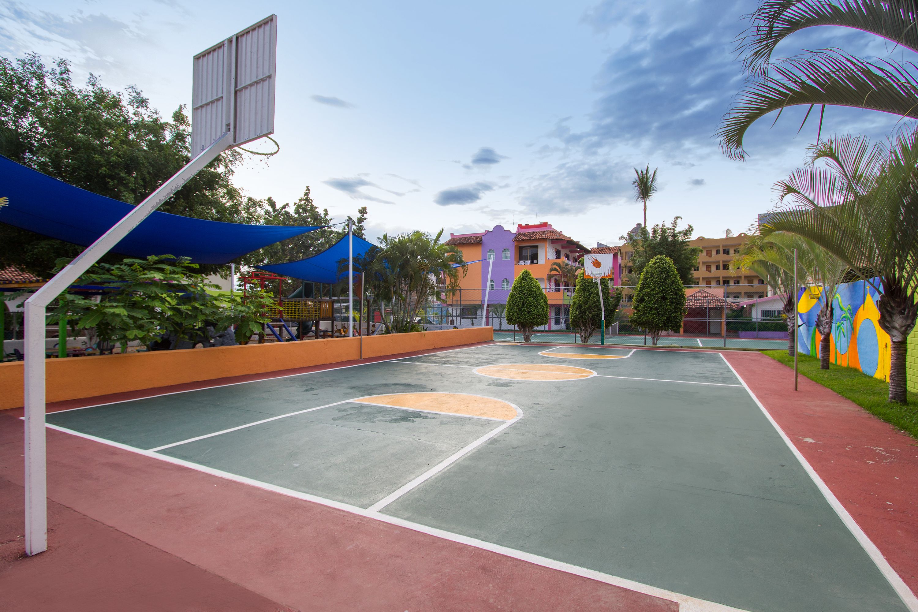 An outdoor basketball court with a blue canopy, surrounded by palm trees and colorful buildings.