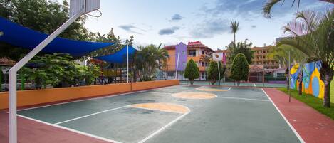 An outdoor basketball court with a blue canopy, surrounded by palm trees and colorful buildings.