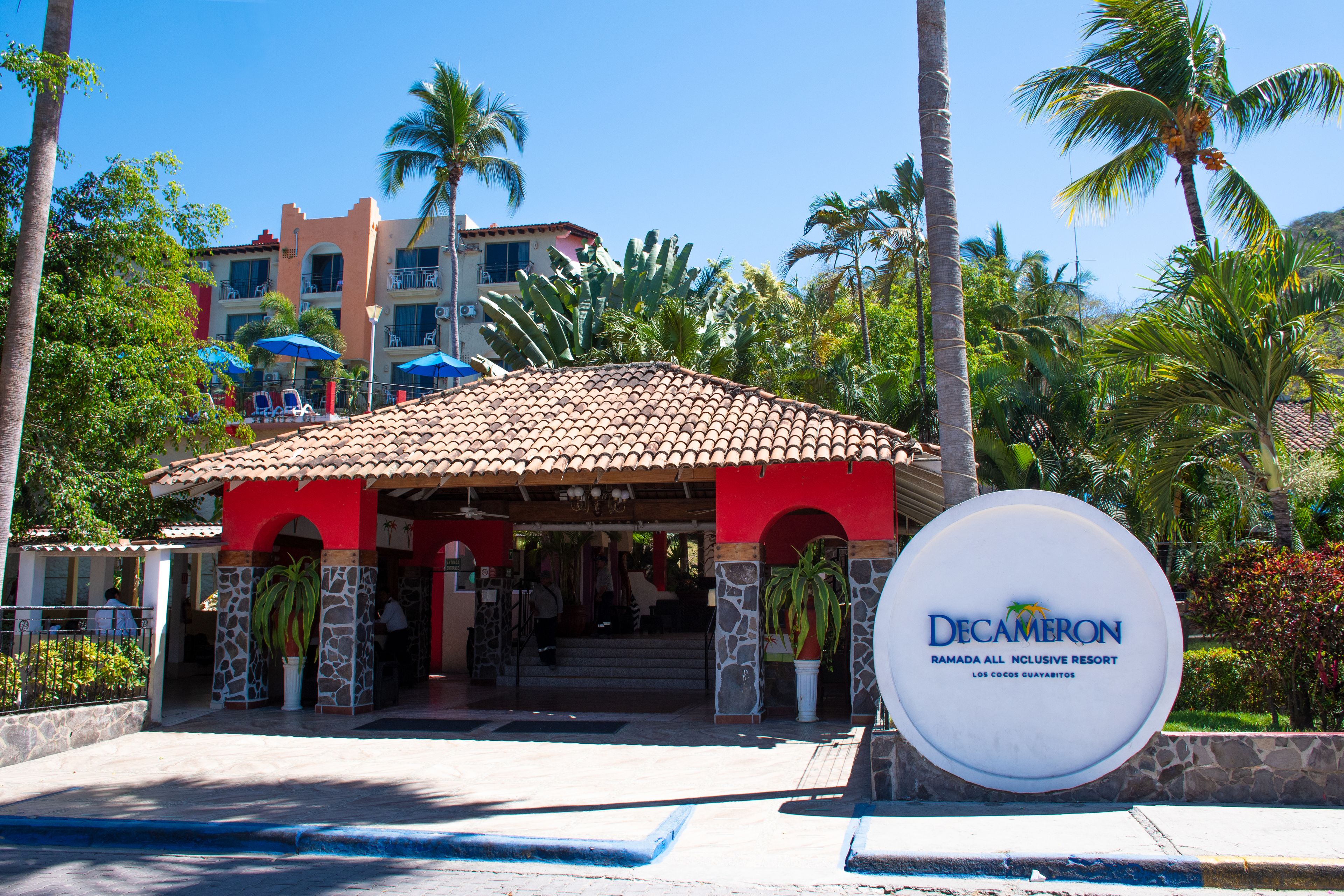 Entrada a un resort con un edificio rojo y blanco, palmeras y un letrero de Decameron.