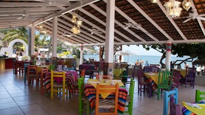 Un área de comedor al aire libre con sillas y mesas de colores, piso de baldosas y vista al mar.