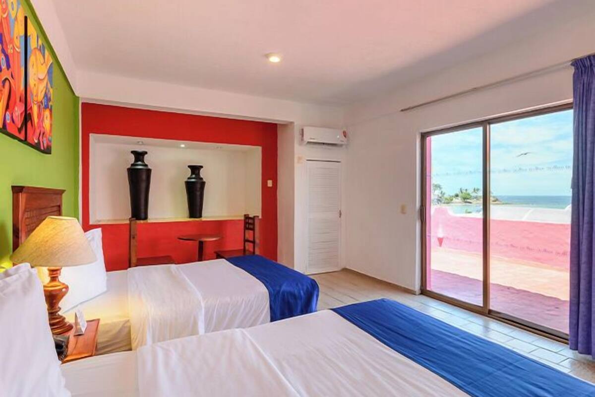A hotel room with two beds, a desk, a chair, a lamp, a mirror, and a view of the beach.