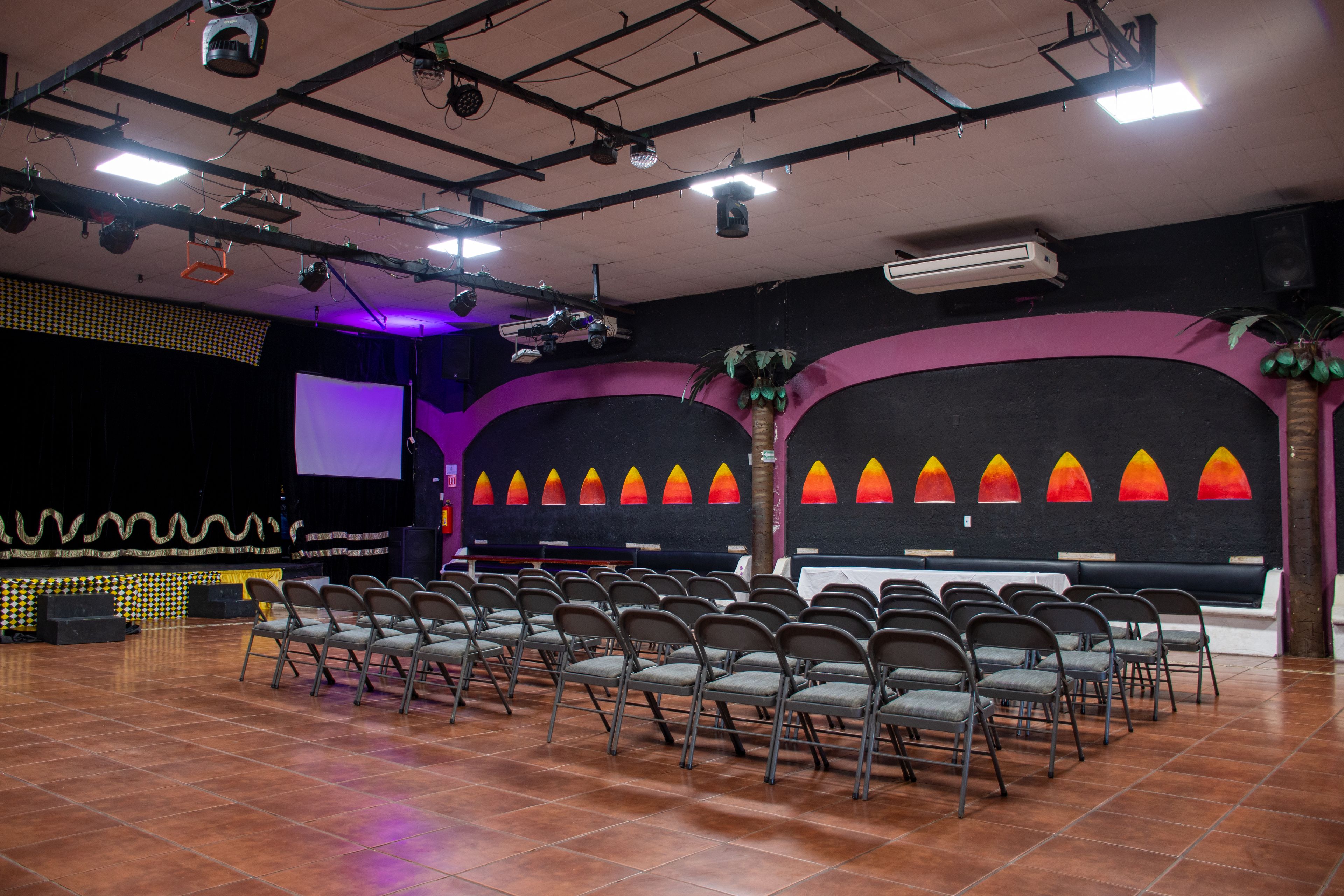 Un auditorio con hileras de sillas, un escenario con una pantalla grande e iluminación en el techo.