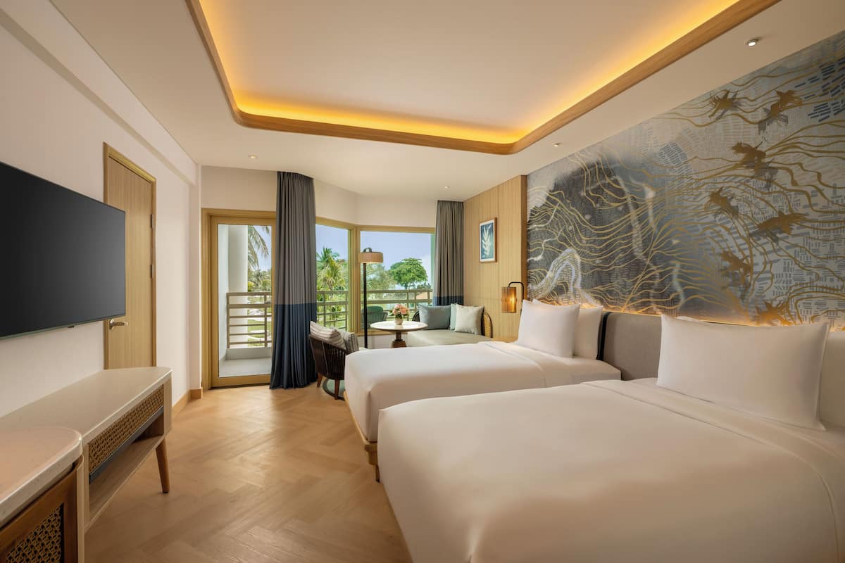 A hotel room with two beds, a TV, a desk, and a balcony with a view of palm trees.