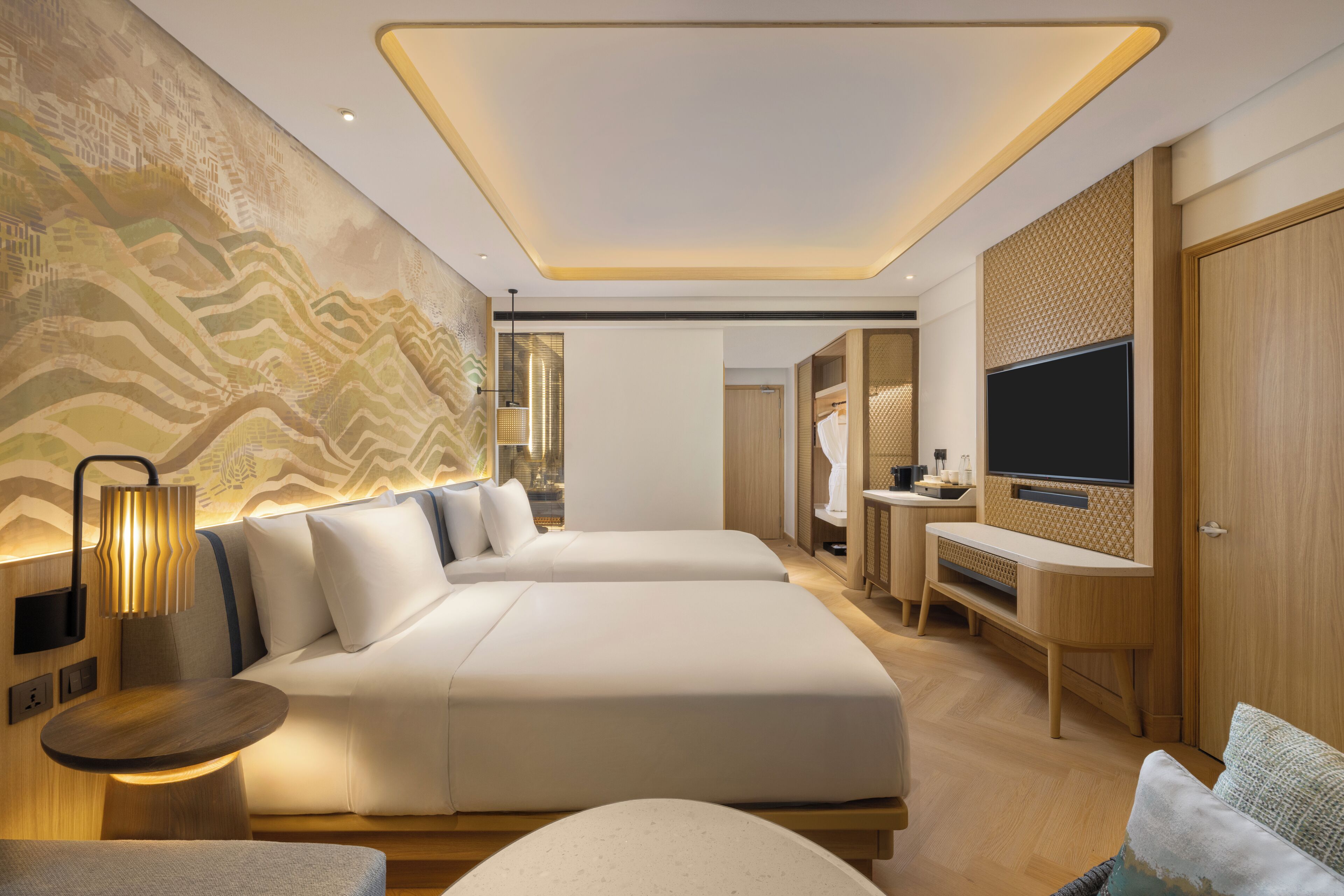 A modern hotel room with two beds, a TV, a desk, and a bathroom visible in the background.