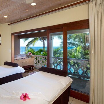 A room with two beds, a ceiling fan, a view of the ocean, and a balcony with a wooden railing.