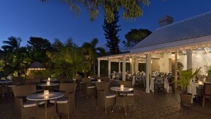 An outdoor patio area with round tables and wicker chairs, surrounded by palm trees and a covered seating area with string lights.
