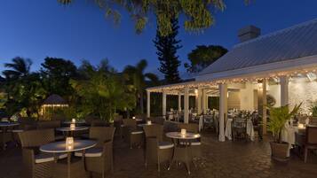 An outdoor patio area with round tables and wicker chairs, surrounded by palm trees and a covered seating area with string lights.