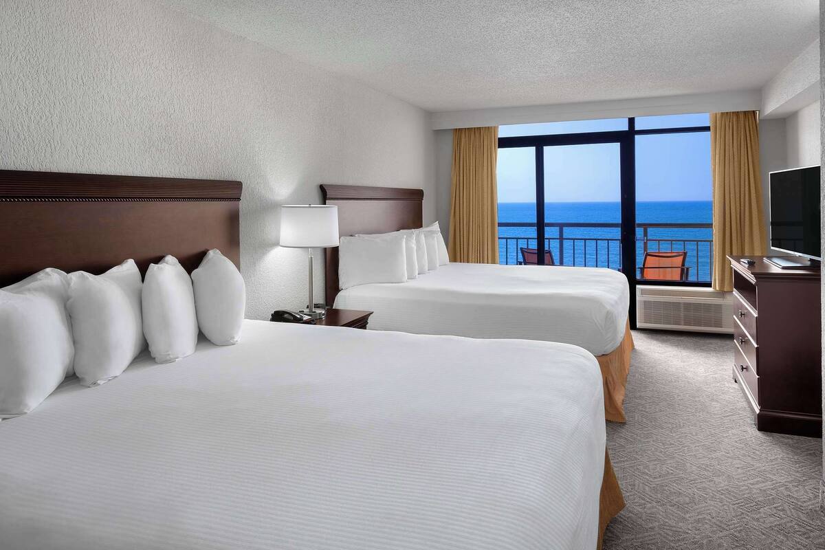 Habitación de hotel con dos camas, un televisor y un ventanal con vistas al océano.