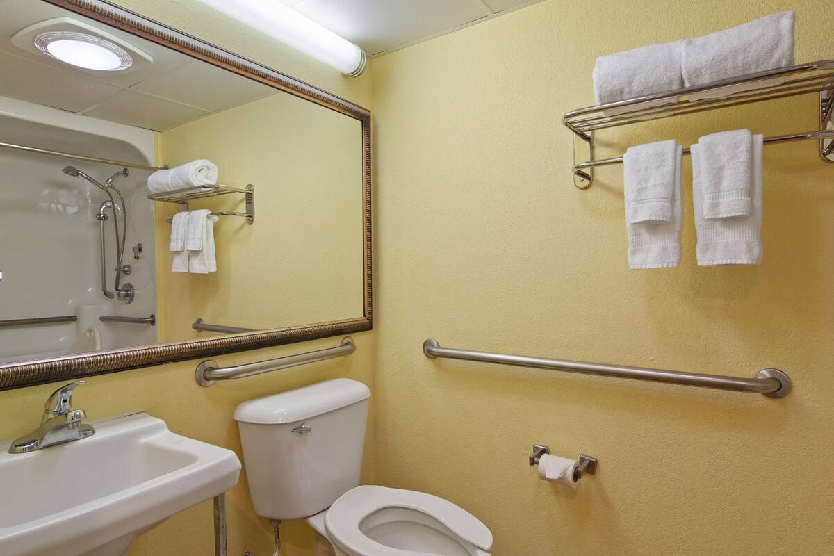 Un baño con lavamanos, inodoro y espejo. Hay barras de apoyo en las paredes y un perchero con toallas.