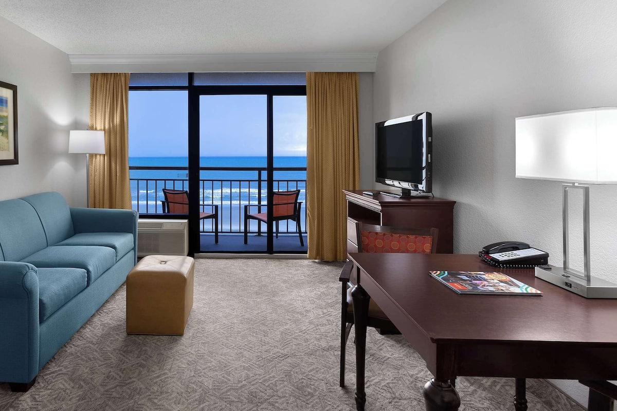 Habitación de hotel con sofá azul, una mesa de madera, televisión y vista al océano.
