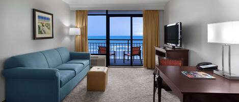 A hotel room with a blue sofa, a wooden table, a TV, and a view of the ocean.