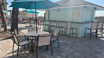 Un patio al aire libre con mesas y sillas de metal, una zona de estar cubierta con sombrillas y un pequeño edificio con un letrero.