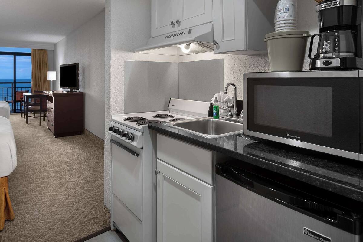 Habitación de hotel con cama, escritorio, televisor y cocina equipada con estufa, microondas y lavaplatos.