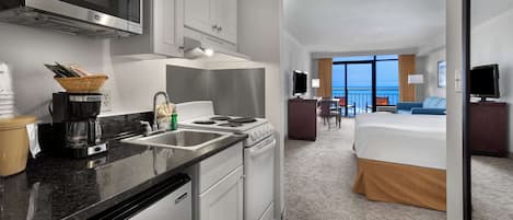 A hotel room with a kitchenette, a bed, a TV, and a view of the ocean.