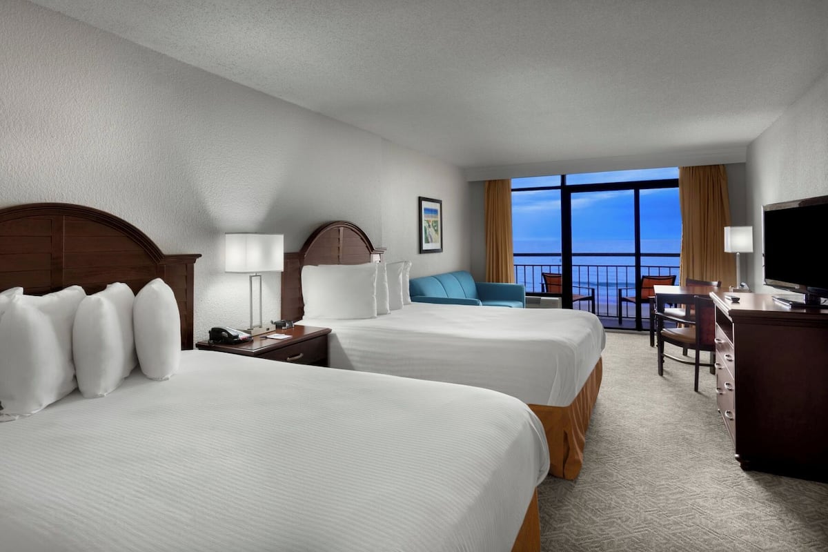 Habitación de hotel con dos camas, un escritorio, un televisor y un balcón con vistas.