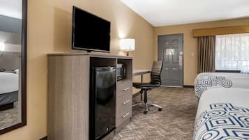 A hotel room with a flat-screen TV mounted on a wooden cabinet, a desk with a chair, a bed with patterned bedding, and a door with a window view.