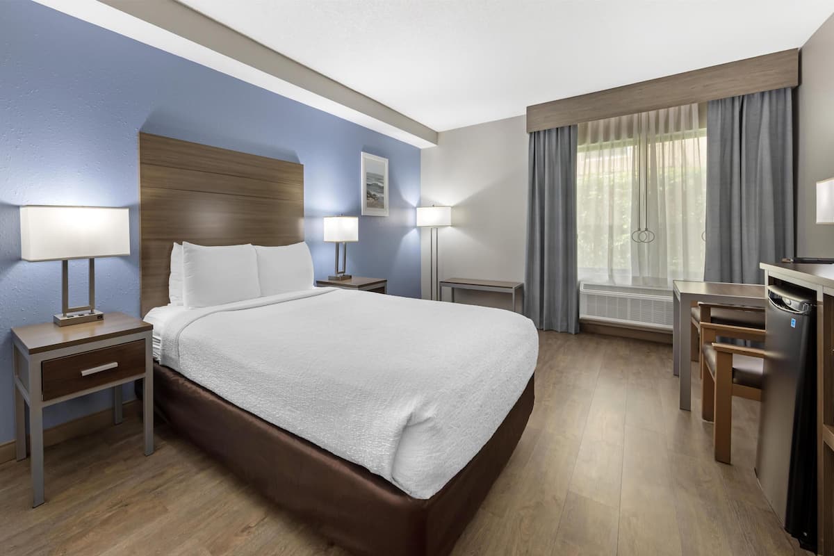 A hotel room with a large bed, bedside tables with lamps, a desk with a chair, a window with curtains, and a view of greenery outside.