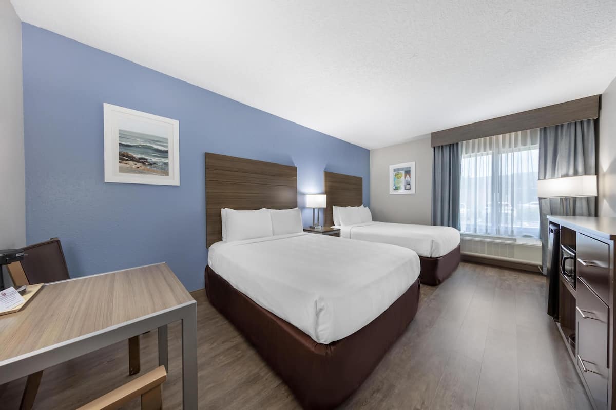 A hotel room with two beds, a desk, a chair, a desk lamp, a nightstand, a window with curtains, and a wall-mounted TV.