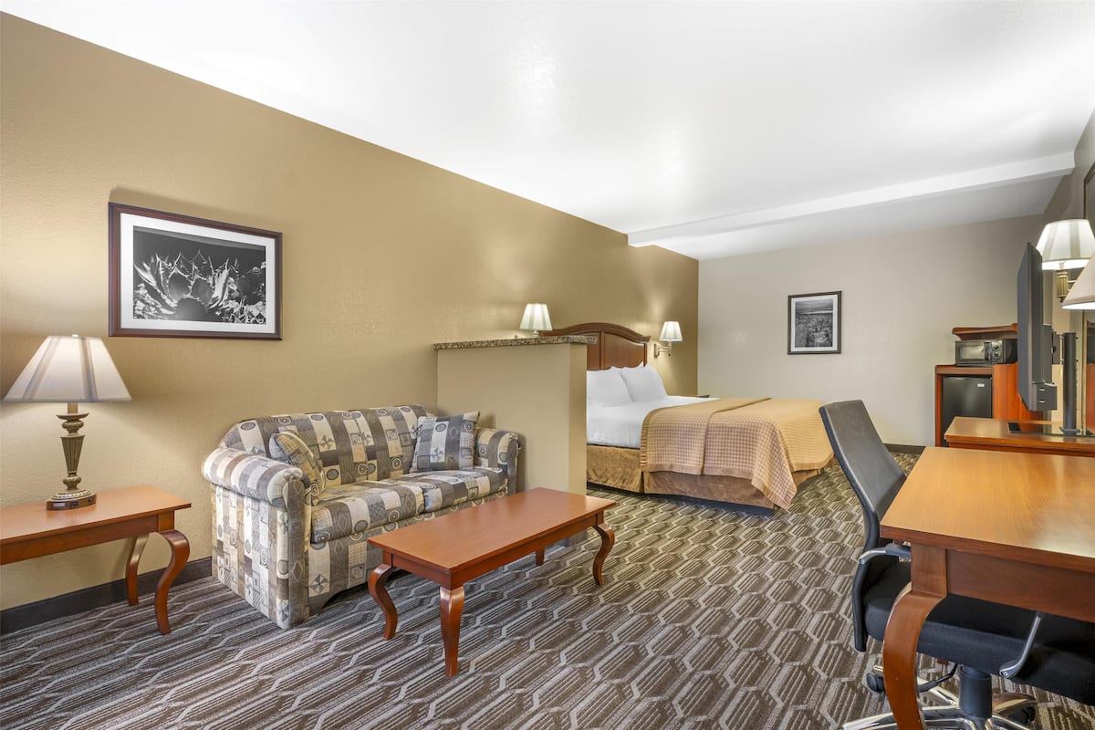 A hotel room with a patterned sofa, a wooden table, a bed with a white comforter, and a desk with a lamp.