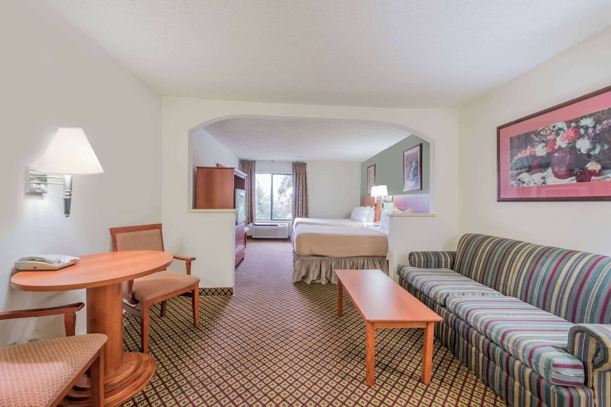 A hotel room with a bed, a striped sofa, a table, and a chair. There is a window with curtains, a lamp, and a framed picture on the wall.